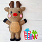 Kawaii amigurumi ragdoll style crochet pattern for a reindeer toy with antlers, red nose, and pink cheeks on a light wooden background with colorful gift boxes.