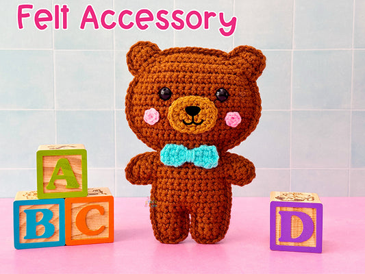 Felt nose accessories for a crocheted brown bear with a blue bow tie standing next to wooden blocks on a pink surface.