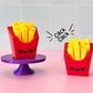 Two red fidget clickers with yellow french fries on a pink surface with a white tiled background.