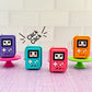 Four colorful handheld gaming console-shaped clicker keychains on a light purple surface with a white tiled background.