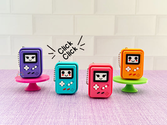Four colorful handheld gaming console-shaped clicker keychains on a light purple surface with a white tiled background.