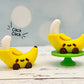 Two banana-shaped fidget clickers with faces on a wooden surface, one on a small green stand.
