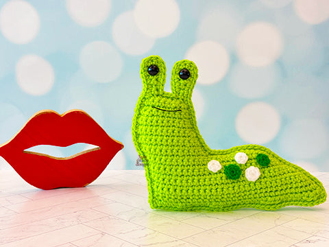 Kawaii amigurumi ragdoll style crochet pattern for a Green crocheted slug toy next to a pair of red lips on a light blue background with white circles.