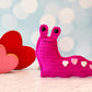 Kawaii amigurumi ragdoll style crochet pattern for a pink slug toy with heart patterns next to red hearts on a light blue background