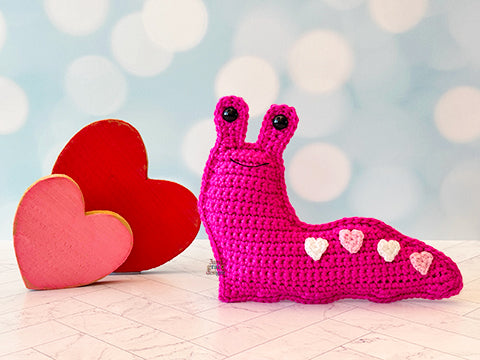 Kawaii amigurumi ragdoll style crochet pattern for a pink slug toy with heart patterns next to red hearts on a light blue background
