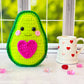 A kawaii amigurumi ragdoll style crochet pattern for a avocado with a pink heart on a windowsill with a mug and heart decorations.