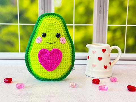 A kawaii amigurumi ragdoll style crochet pattern for a avocado with a pink heart on a windowsill with a mug and heart decorations.