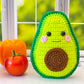 A kawaii amigurumi ragdoll style crochet pattern for a avocado toy with a happy face next to real vegetables on a windowsill.