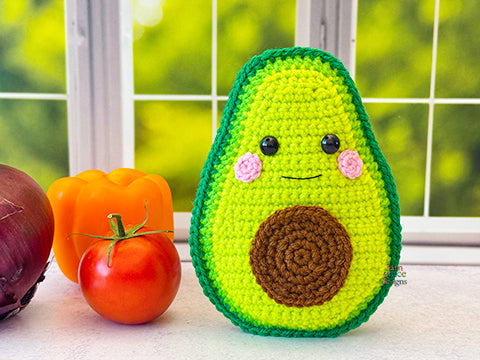 A kawaii amigurumi ragdoll style crochet pattern for a avocado toy with a happy face next to real vegetables on a windowsill.