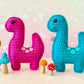 A kawaii amigurumi ragdoll style crochet pattern for a brontosaurus dinosaur. Pictured are two crocheted dinosaur toys, one pink and one blue, with colorful mushrooms on a light background.