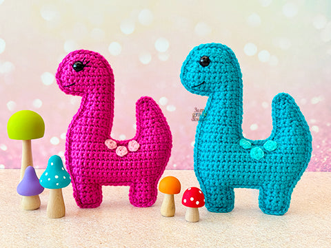 A kawaii amigurumi ragdoll style crochet pattern for a brontosaurus dinosaur. Pictured are two crocheted dinosaur toys, one pink and one blue, with colorful mushrooms on a light background.
