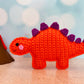 Miniature crocheted stegosaurus standing next to a volcano on a sand surface with a blue and white background