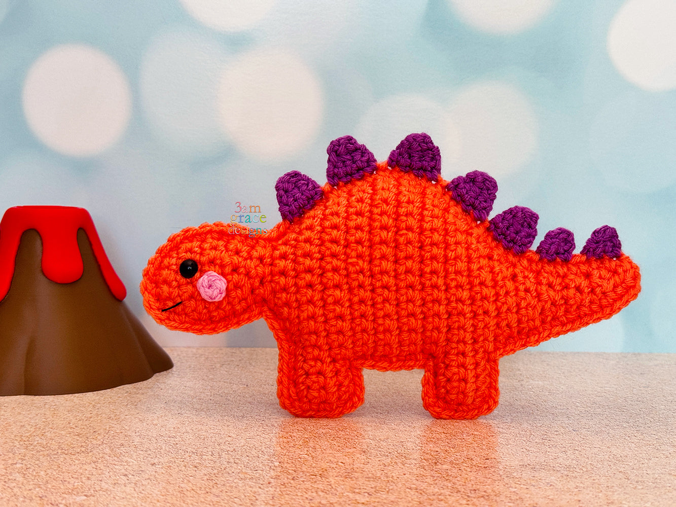 Miniature crocheted stegosaurus standing next to a volcano on a sand surface with a blue and white background