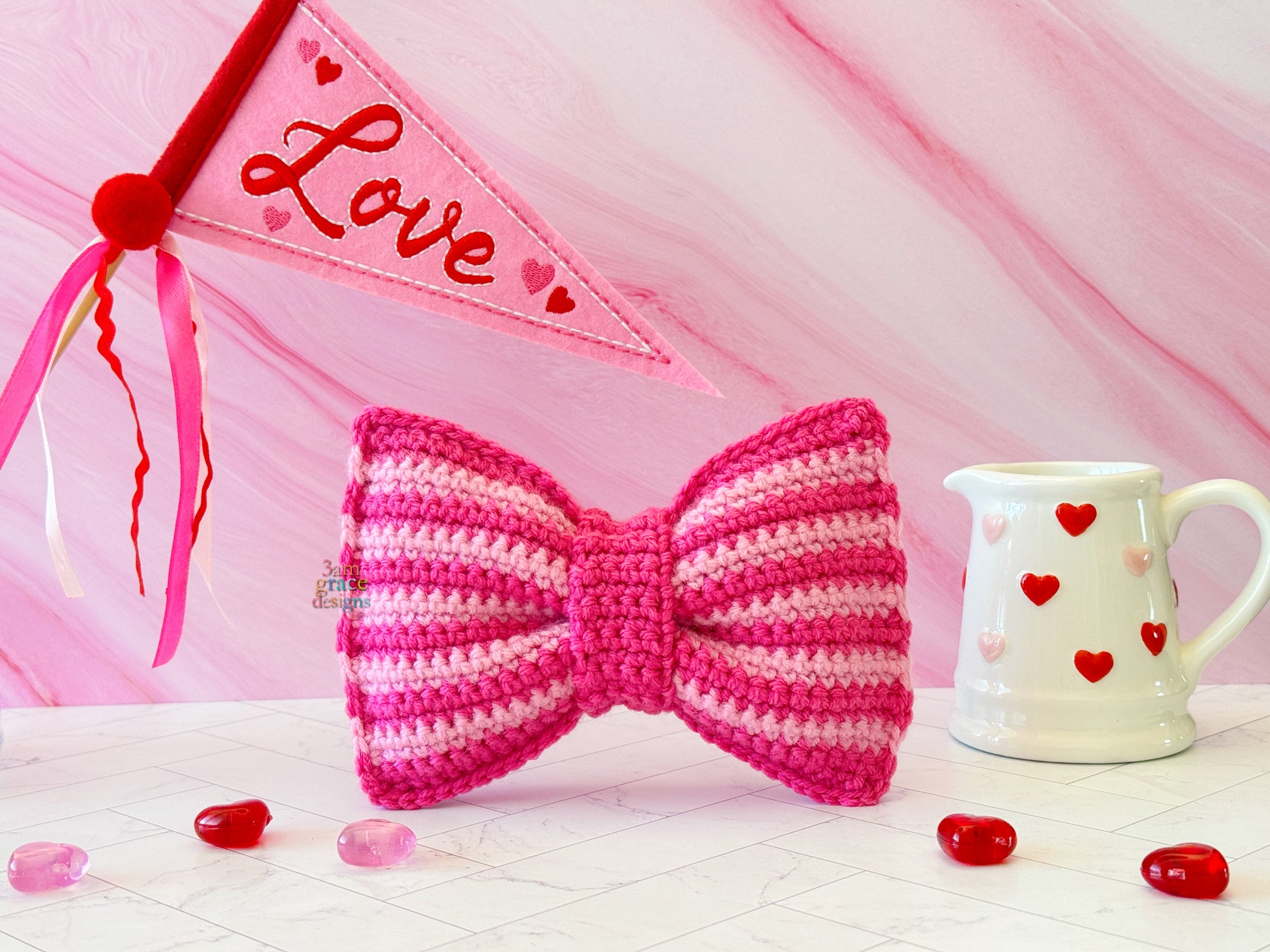 Kawaii amigurumi ragdoll style crochet pattern for a pink bow with a 'Love' banner and heart-shaped decorations on a pink background