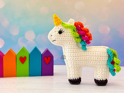 Kawaii amigurumi ragdoll style crochet pattern for a unicorn toy with a colorful mane and tail in front of rainbow-colored houses.
