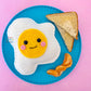 Crocheted egg on a turquoise plate next to toast & orange slices on a pink background.