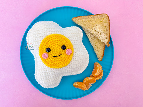 Crocheted egg on a turquoise plate next to toast & orange slices on a pink background.
