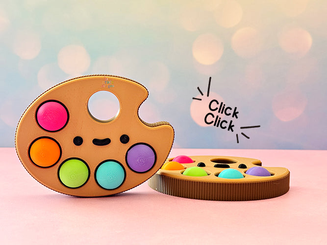 Colorful paint palette with kawaii face fidget clicker and 'Click Click' text on a blurred background