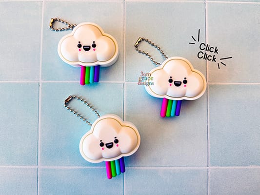 Three cloud-shaped fidget clicker keychains with rainbow tails on a tiled background