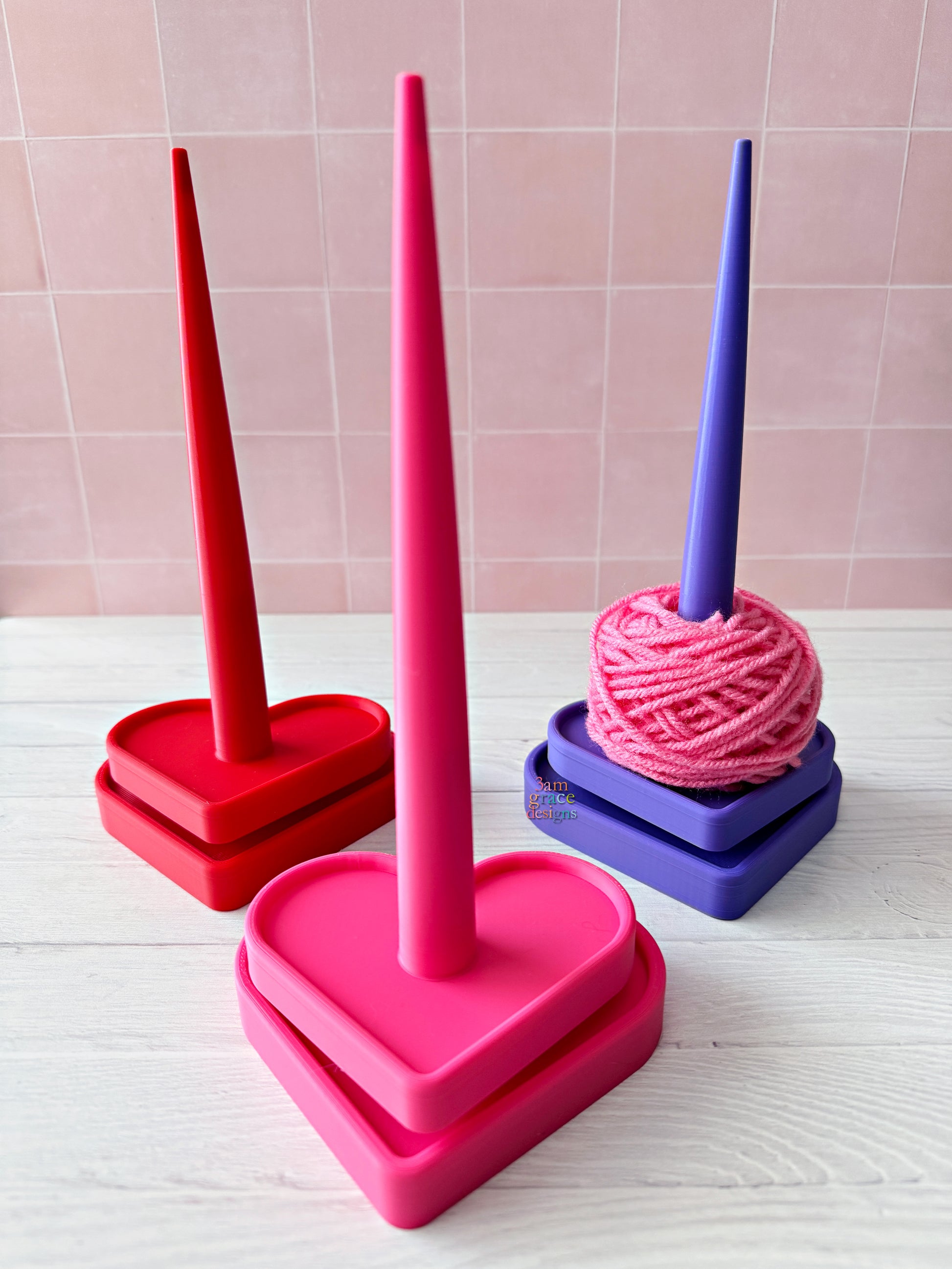3d printed yarn spinner tool in shape of heart in bright colors. Pictured on a light wooden surface with a pink tiled wall background