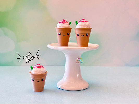 Three small frappucino fidget clickers with faces on a white stand against a blurred background.