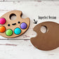 Colorful paint palette with kawaii face fidget clicker labeled 'Imperfect Version'.