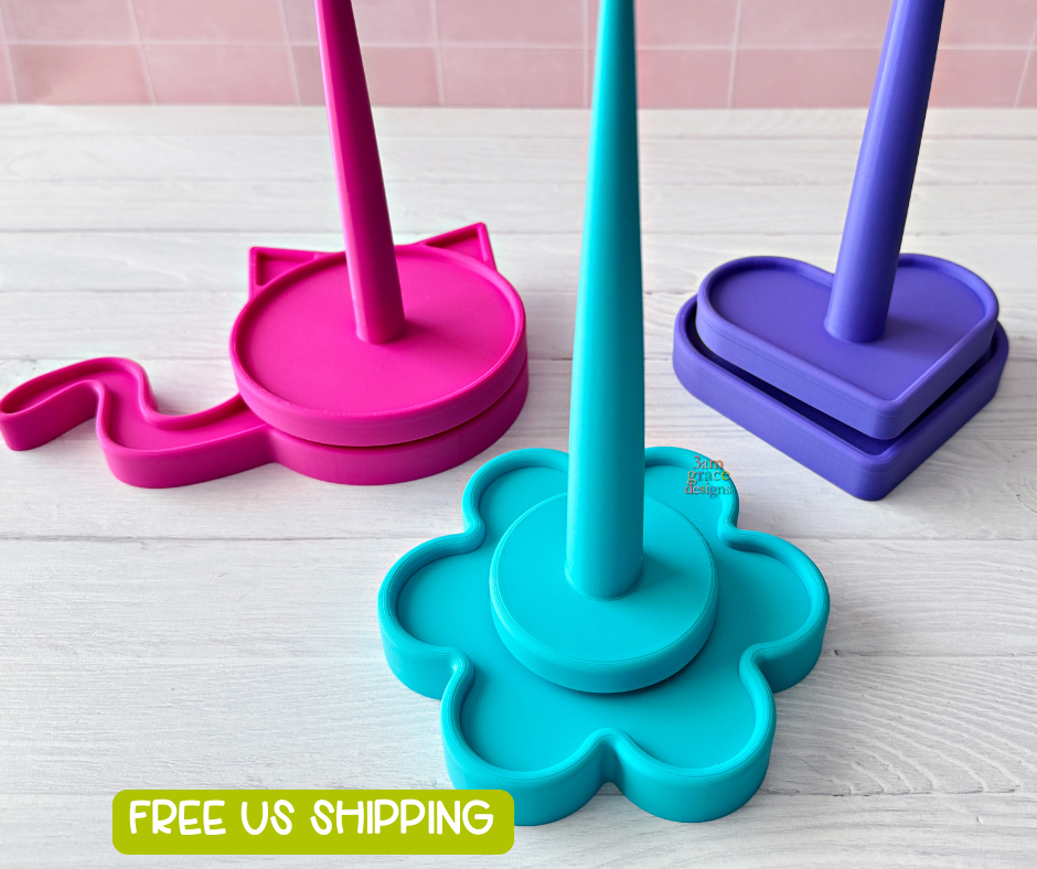 3d printed yarn spinner tool in shape of heart, flower, and cat in bright colors. Pictured on a light wooden surface