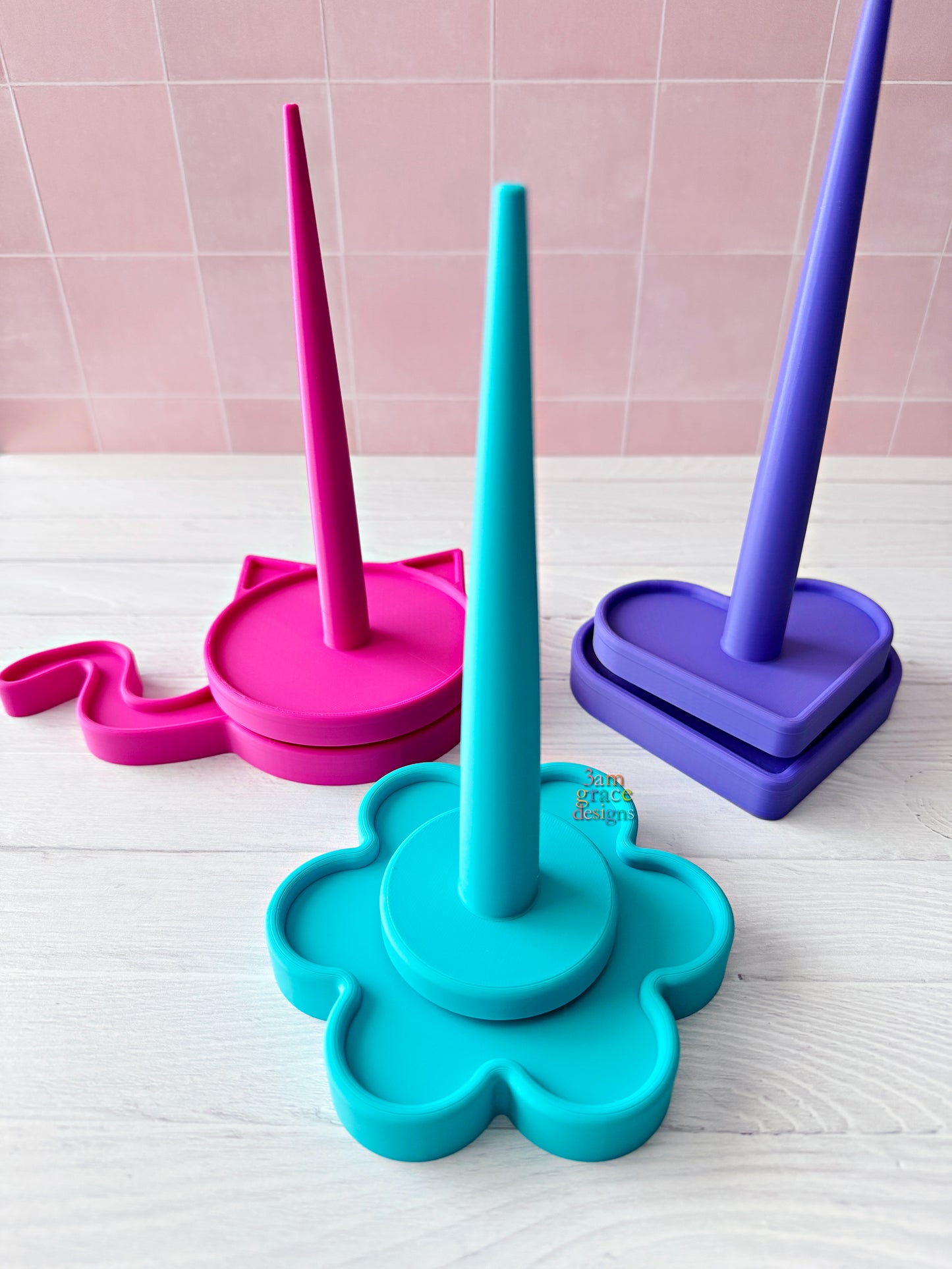 3d printed yarn spinner tool in shape of heart, flower, and cat in bright colors. Pictured on a white surface with a pink tiled wall background